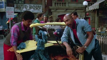 Movie still from “Follow That Bird” (1985), directed by Ken Kwapis – A group of people standing next to a sesame street car; Medium shot, Over the shoulder angle