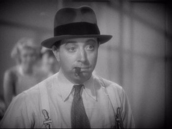 Movie still from “Footlight Parade” (1933), directed by Lloyd Bacon – A man in a hat smoking a cigarette in a black and white photo; Close Up shot, Low angle