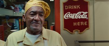 Movie still from “Footloose” (2011), directed by Craig Brewer – An older man wearing a yellow hat standing in front of a coca - cola sign; Close Up shot, Low angle
