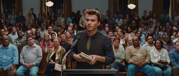 Movie still from “Footloose” (2011), directed by Craig Brewer – A man standing at a podium holding a book in front of an audience; Medium shot, Over the shoulder angle