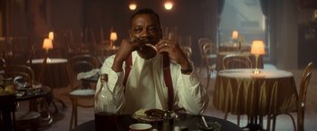 Movie still from “The United States vs. Billie Holiday” (2021), directed by Lee Daniels – A man sitting at a table eating a doughnut; Medium shot, Low angle