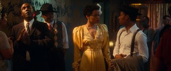 Movie still from “The United States vs. Billie Holiday” (2021), directed by Lee Daniels – A woman in a yellow dress standing next to two other men; Medium shot, Over the shoulder angle