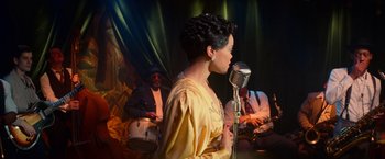 Movie still from “The United States vs. Billie Holiday” (2021), directed by Lee Daniels – A woman is singing into a microphone while a man plays drums behind her; Medium shot, Over the shoulder angle