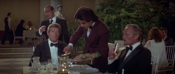 Movie still from “For Your Eyes Only” (1981), directed by John Glen – A man holding a plate of food at a dinner table; Medium shot, Over the shoulder angle