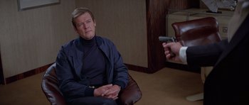 Movie still from “For Your Eyes Only” (1981), directed by John Glen – A man sitting in a chair with a gun pointed at him; Medium shot, Over the shoulder angle