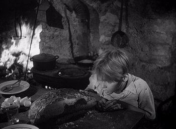 Movie still from “Forbidden Games” (1952), directed by René Clément – A young boy eating a loaf of bread in a kitchen; Medium shot, Over the shoulder angle