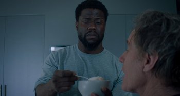 Movie still from “The Upside” (2017), directed by Neil Burger – A man eating a bowl of cereal while another man watches; Medium shot, Over the shoulder angle
