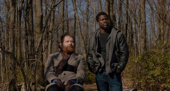Movie still from “The Upside” (2017), directed by Neil Burger – Two men are standing in the woods and one is sitting on the ground and the other is standing up; Medium shot, Over the shoulder angle