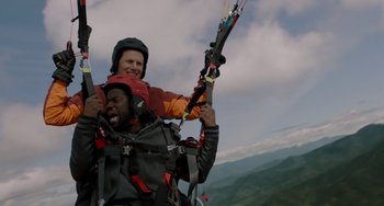Movie still from “The Upside” (2017), directed by Neil Burger – A man holding onto the back of another man who is holding onto a paraglider; Medium shot, Low angle