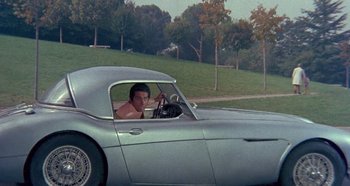 Movie still from “Four Times That Night” (1971), directed by Mario Bava – A young man driving a silver sports car in a field; Wide shot, High angle