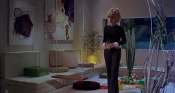 Movie still from “Four Times That Night” (1971), directed by Mario Bava – A woman standing in a living room next to a robot; Wide shot, Low angle