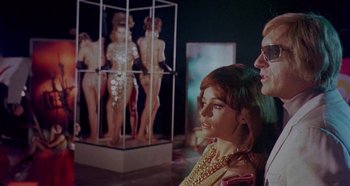 Movie still from “Four Times That Night” (1971), directed by Mario Bava – A woman and a man standing next to each other in front of a display case; Medium shot, Low angle