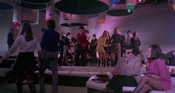 Movie still from “Four Times That Night” (1971), directed by Mario Bava – A group of people standing around in a room; Wide shot, Low angle