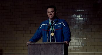 Movie still from “Foxcatcher” (2014), directed by Bennett Miller – A man standing at a podium with a medal around his neck; Medium shot, Over the shoulder angle