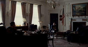 Movie still from “Foxcatcher” (2014), directed by Bennett Miller – A man sitting on a chair in front of an american flag; Wide shot, Over the shoulder angle