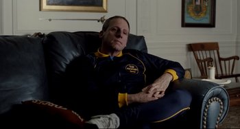 Movie still from “Foxcatcher” (2014), directed by Bennett Miller – A man sitting in a chair with his hands folded on his chest; Medium shot, Over the shoulder angle