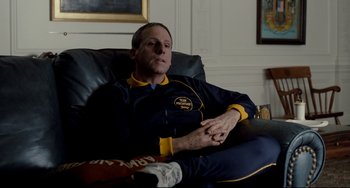 Movie still from “Foxcatcher” (2014), directed by Bennett Miller – A man sitting in a chair in a room; Medium shot, Over the shoulder angle