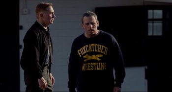 Movie still from “Foxcatcher” (2014), directed by Bennett Miller – Two men standing next to each other in front of a building; Medium shot, Over the shoulder angle
