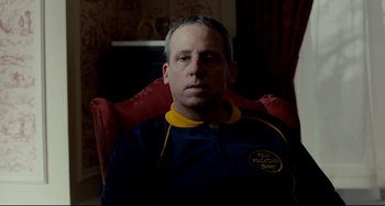 Movie still from “Foxcatcher” (2014), directed by Bennett Miller – A man sitting in a red chair wearing a blue and yellow jacket; Close Up shot, Over the shoulder angle