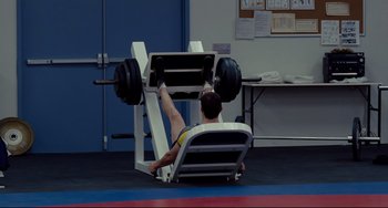 Movie still from “Foxcatcher” (2014), directed by Bennett Miller – A man sitting in a chair lifting a barbell; Wide shot, High angle