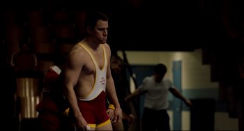 Movie still from “Foxcatcher” (2014), directed by Bennett Miller – A man standing in a wrestling uniform in a gym; Medium shot, Over the shoulder angle