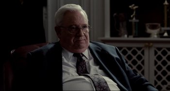 Movie still from “Foxcatcher” (2014), directed by Bennett Miller – An older man wearing a suit and tie sitting in a chair; Medium shot, Over the shoulder angle