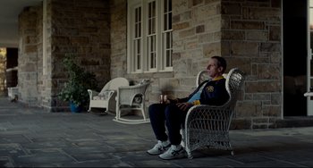 Movie still from “Foxcatcher” (2014), directed by Bennett Miller – A man sitting on a chair outside of a house; Medium shot, High angle