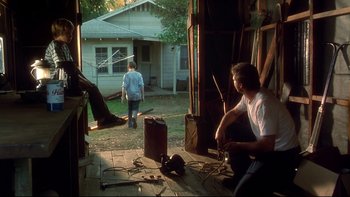 Movie still from “Frailty” (2001), directed by Bill Paxton – A man sitting on the porch of a house while another man walks by; Wide shot, Over the shoulder angle