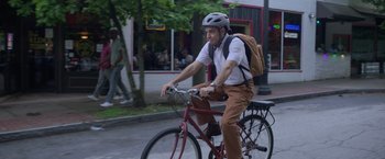Movie still from “The Valet” (2022), directed by Richard Wong – A man riding a bike down a street with trees in the background; Medium shot, Low angle