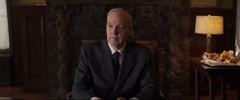 Movie still from “French Exit” (2020), directed by Azazel Jacobs – An older man wearing a suit and tie sitting in a chair in front of an ornate wall; Medium shot, Over the shoulder angle