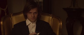 Movie still from “French Exit” (2020), directed by Azazel Jacobs – A man sitting in a chair holding a glass; Close Up shot, Low angle