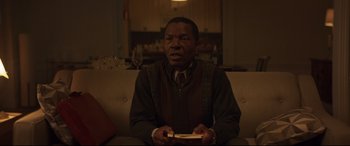 Movie still from “French Exit” (2020), directed by Azazel Jacobs – A man sitting on top of a couch holding a nintendo wii game controller; Medium shot, Over the shoulder angle