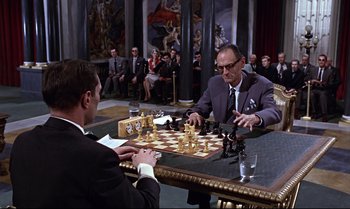 Movie still from “From Russia with Love” (1963), directed by Terence Young – Two men in suits playing a game of chess in front of a crowd of onlookers; Medium shot, Over the shoulder angle