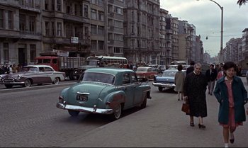 Movie still from “From Russia with Love” (1963), directed by Terence Young – An old car is driving down a busy street; Wide shot, High angle