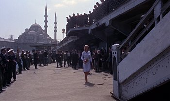 Movie still from “From Russia with Love” (1963), directed by Terence Young – A woman walking down a street near a crowd of onlookers; Extreme Wide shot, Low angle