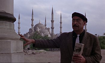 Movie still from “From Russia with Love” (1963), directed by Terence Young – A man holding a book in front of a building; Medium shot, Low angle