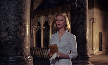 Movie still from “From Russia with Love” (1963), directed by Terence Young – A woman holding a book while standing next to a pillar; Medium shot, Low angle
