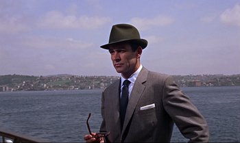 Movie still from “From Russia with Love” (1963), directed by Terence Young – A man in a suit and hat standing in front of a body of water; Medium shot, Low angle