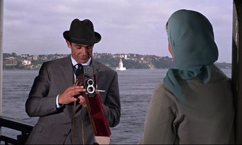 Movie still from “From Russia with Love” (1963), directed by Terence Young – A man in a suit and hat taking a picture of a woman in front of a body of water; Medium shot, Over the shoulder angle