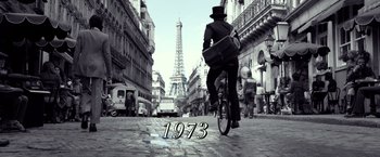 Movie still from “The Walk” (2015), directed by Robert Zemeckis – A man riding a bike down a street with the eiffel tower in the background; Wide shot, Low angle