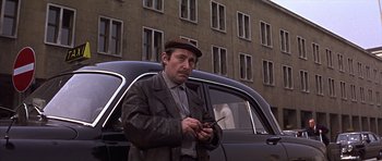 Movie still from “Funeral in Berlin” (1966), directed by Guy Hamilton – A man standing in front of a car holding a cell phone; Medium shot, Low angle