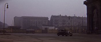 Movie still from “Funeral in Berlin” (1966), directed by Guy Hamilton – An old car driving down a street past a building; Extreme Wide shot, Over the shoulder angle