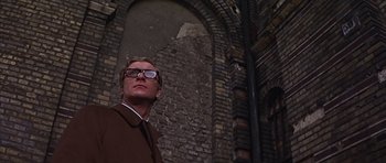 Movie still from “Funeral in Berlin” (1966), directed by Guy Hamilton – A man wearing glasses standing in front of a brick wall; Close Up shot, Low angle