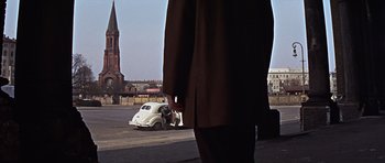 Movie still from “Funeral in Berlin” (1966), directed by Guy Hamilton – A man standing in front of an old white car; Wide shot, Low angle