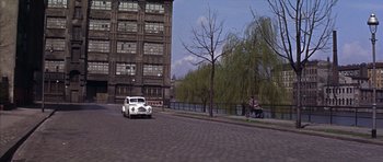 Movie still from “Funeral in Berlin” (1966), directed by Guy Hamilton – An old white car driving down a street; Extreme Wide shot, High angle