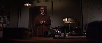 Movie still from “Funeral in Berlin” (1966), directed by Guy Hamilton – A man in a brown coat standing in front of a table; Medium shot, Low angle