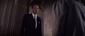 Movie still from “Funeral in Berlin” (1966), directed by Guy Hamilton – A man wearing a suit and tie standing next to a wall; Medium shot, Low angle