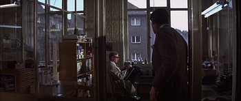 Movie still from “Funeral in Berlin” (1966), directed by Guy Hamilton – A man sitting in a chair in front of a large window; Wide shot, Over the shoulder angle