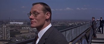 Movie still from “Funeral in Berlin” (1966), directed by Guy Hamilton – A man in a suit and tie standing on a balcony; Close Up shot, Low angle
