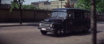Movie still from “Funeral in Berlin” (1966), directed by Guy Hamilton – An old black truck driving down a street; Wide shot, High angle